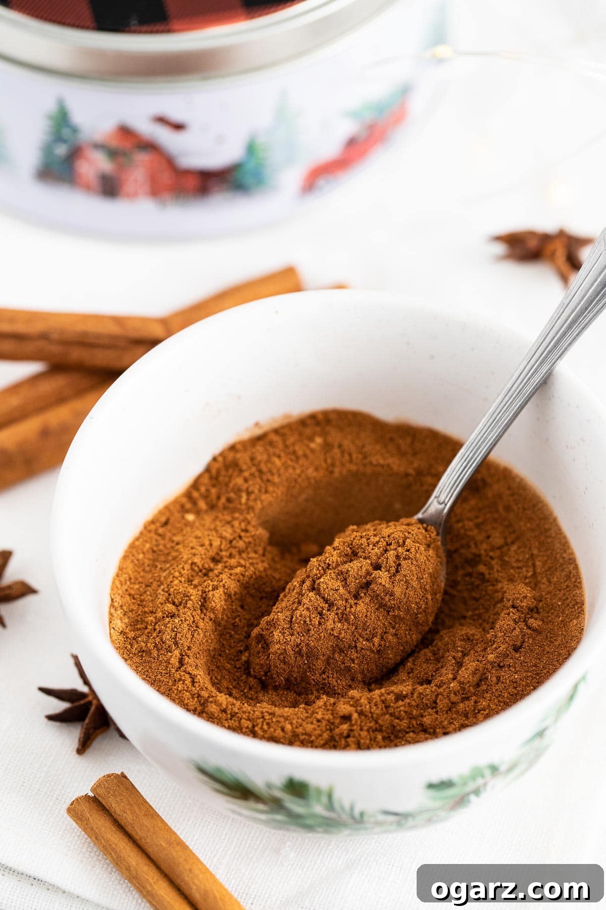 Yuletide Gingerbread Blend 2 A white bowl filled with ground spices and a spoon in it, next to cinnamon sticks and star anise.