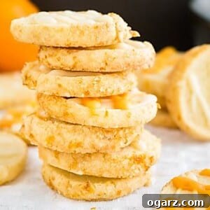 A stack of orange almond cookies on a white tablecloth with more of the cookies next to it and an orange in the background.