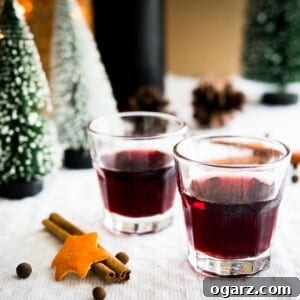 Two glasses of mulled wine liqueur on a grey tablecloth with cinnamon sticks. There are pinecones and little pine trees in the background.
