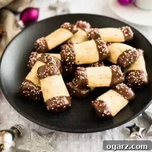 A black plate of maple hazelnut cookies, with both ends dipped in chocolate and sprinkles on a grey tablecloth. There are silver and pink Christmas ornaments lying next to it.