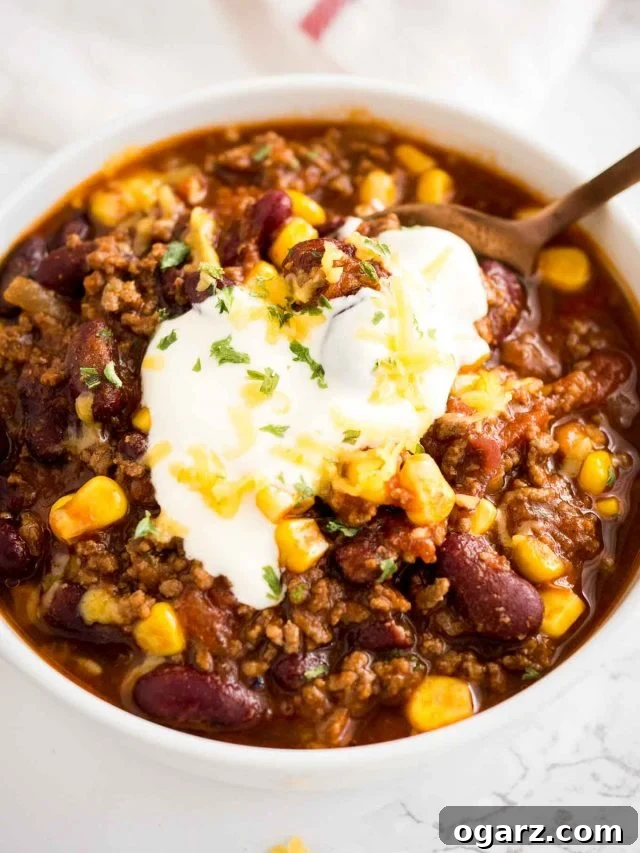 A beautifully presented bowl of Instant Pot Chili con Carne, garnished generously with a dollop of sour cream, fresh green cilantro, and a sprinkle of shredded cheddar cheese. A bronze spoon rests invitingly in the rich, dark red chili, highlighting its inviting texture. The shot is taken from above, showcasing the colorful toppings against the creamy chili.