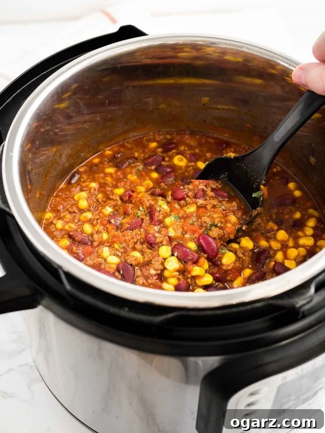 A close-up view inside an Instant Pot, revealing a pot full of rich, simmering chili con carne. A hand holds a black cooking spoon, gently stirring the chili, highlighting its thick texture and vibrant color. The steam suggests its warmth and freshness.