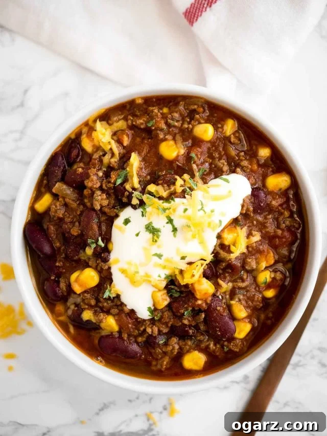 A top-down perspective of a single white bowl of rich chili con carne, adorned with fresh sour cream, bright cilantro, and shredded cheddar cheese. The bowl is set on a light marble surface, accompanied by a neatly folded white towel and an elegant bronze spoon, creating a warm and inviting culinary image.