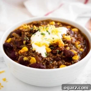 A white bowl of chili con carne, topped with sour cream, cilantro, and shredded cheese, presented on a marble surface alongside a white towel and a bronze spoon. This appetizing image visually represents the delicious Instant Pot Chili recipe.