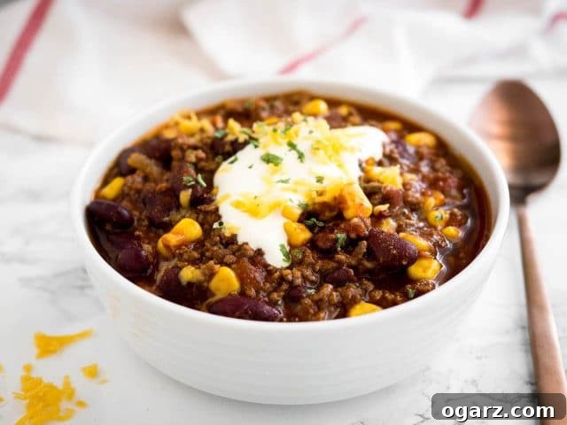 A vibrant, top-down shot of a white bowl filled with Instant Pot Chili con Carne. It is artfully garnished with a generous dollop of sour cream, fresh green cilantro, and a sprinkle of shredded cheddar cheese. The bowl is positioned on a sleek marble surface, with a crisp white towel and an elegant bronze spoon placed beside it, suggesting a comforting and delicious meal.