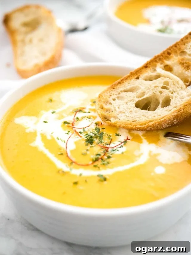 Creamy Instant Pot Carrot Soup 2 A vibrant bowl of creamy Instant Pot Carrot Soup, garnished with a swirl of sour cream and delicate chili threads. A toasted slice of bread and a spoon rest alongside, with a similar bowl and more bread blurred in the background, hinting at a cozy meal setting.