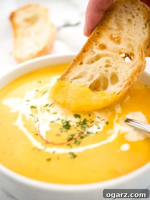 Creamy Instant Pot Carrot Soup 7 A close-up shot of a hand dipping a piece of toasted rustic bread into a bowl of creamy Instant Pot Carrot Soup, garnished with a swirl of sour cream and chili threads. Another slice of bread is visible in the soft background, highlighting the cozy and comforting nature of the meal.