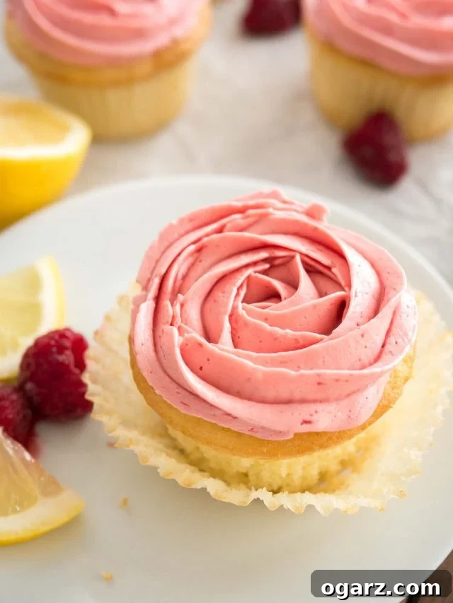 Close-up of a lemon raspberry cupcake with pink raspberry frosting, with the liner folded open on a white plate, garnished with lemon slices and raspberries. There are more of the cupcakes in the background.