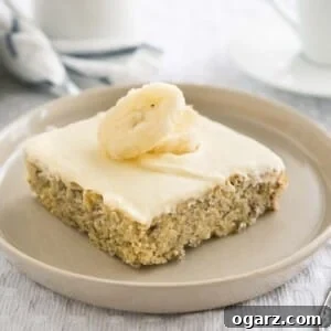 A piece of banana cake with mascarpone frosting and topped with 2 banana slices on a grey plate on a grey tablecloth. There's a fork next to it and a white cup in the background next to a white and blue dishtowel.