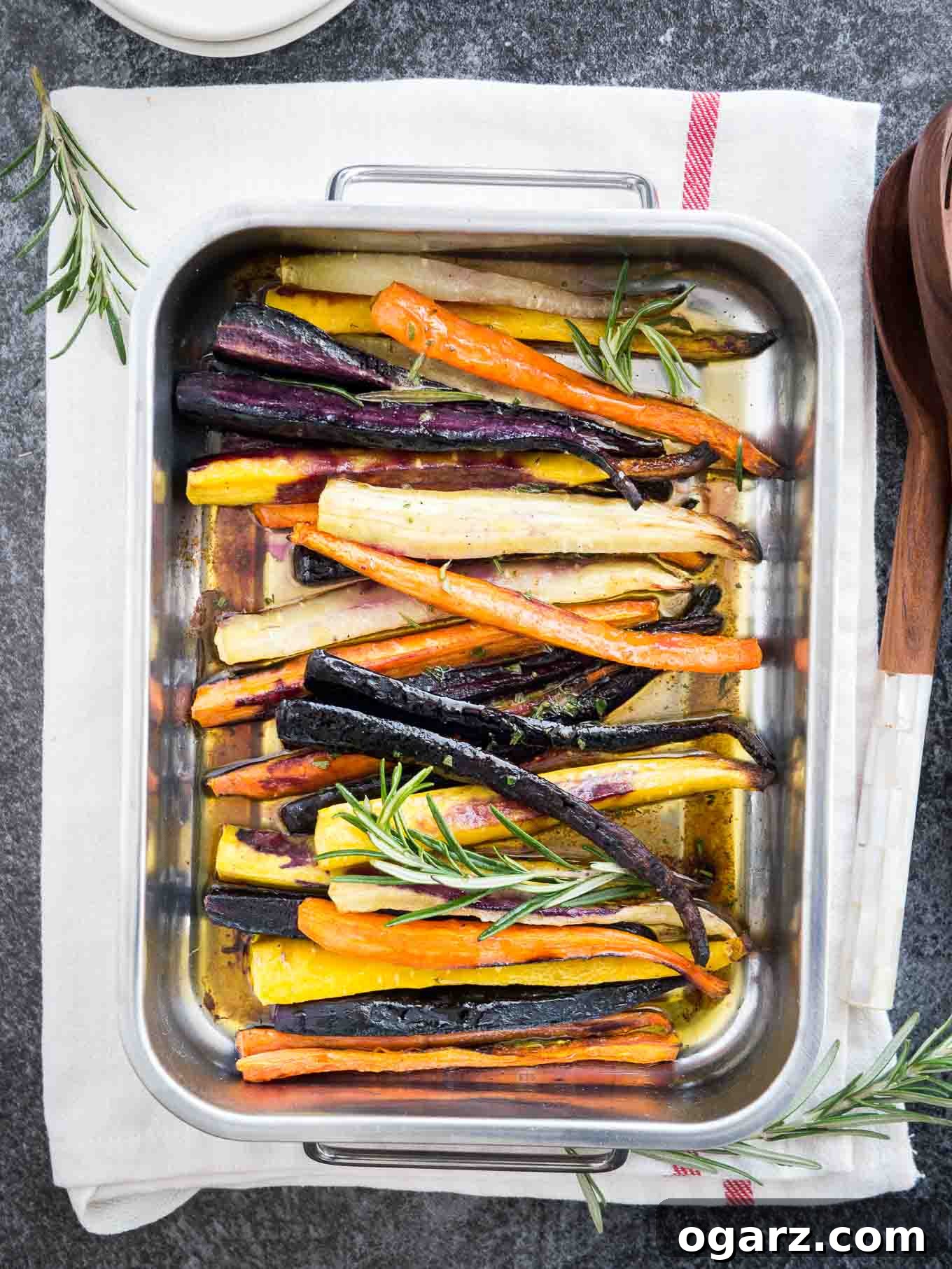Brown Butter Maple Infused Roasted Rainbow Carrots 3 Top-down shot of a roasting pan with roasted rainbow carrots garnished with sprigs of rosemary on a white dishtowel with a red stripe. There are salad tongs next to it.