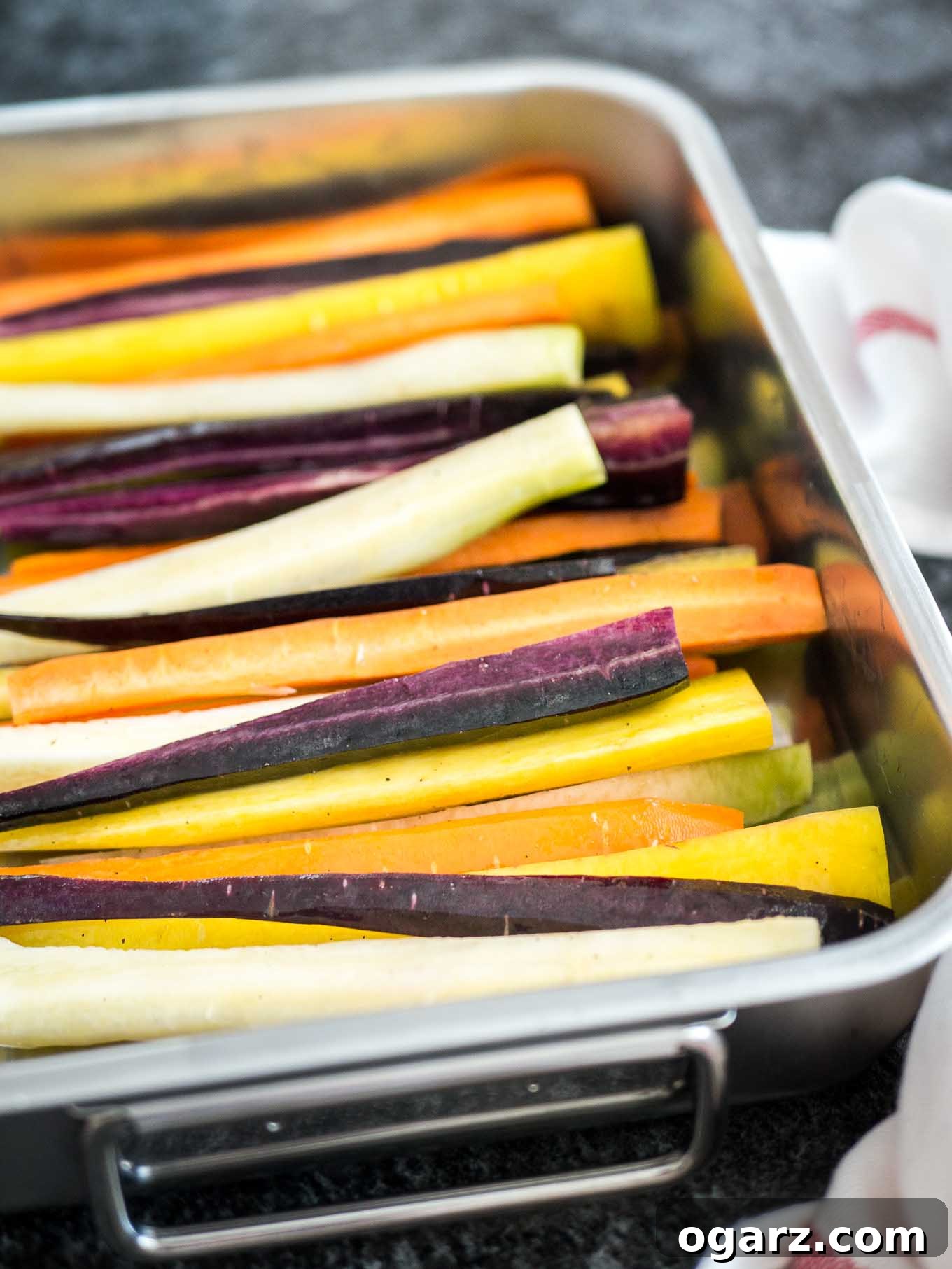Brown Butter Maple Infused Roasted Rainbow Carrots 4 A roasting pan with rainbow carrots cut into wedges lengthwise before baking. There is a white dishtowel with a red stripe next to it.