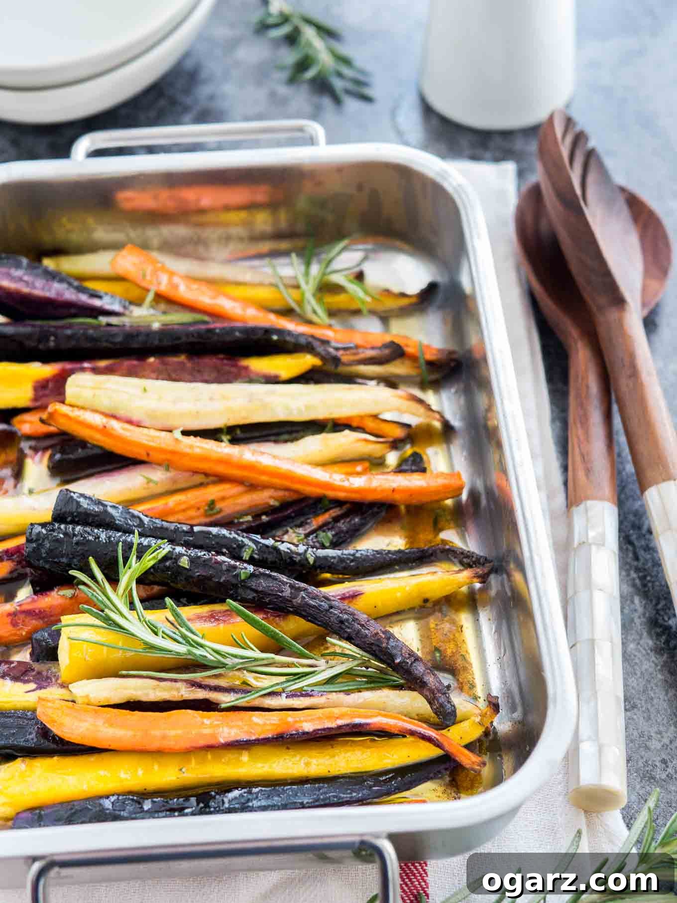 Brown Butter Maple Infused Roasted Rainbow Carrots 5 A roasting pan with roasted rainbow carrots garnished with sprigs of rosemary on a white dishtowel with a red stripe. There is a stack of plates in the background and salad tongs next to it.