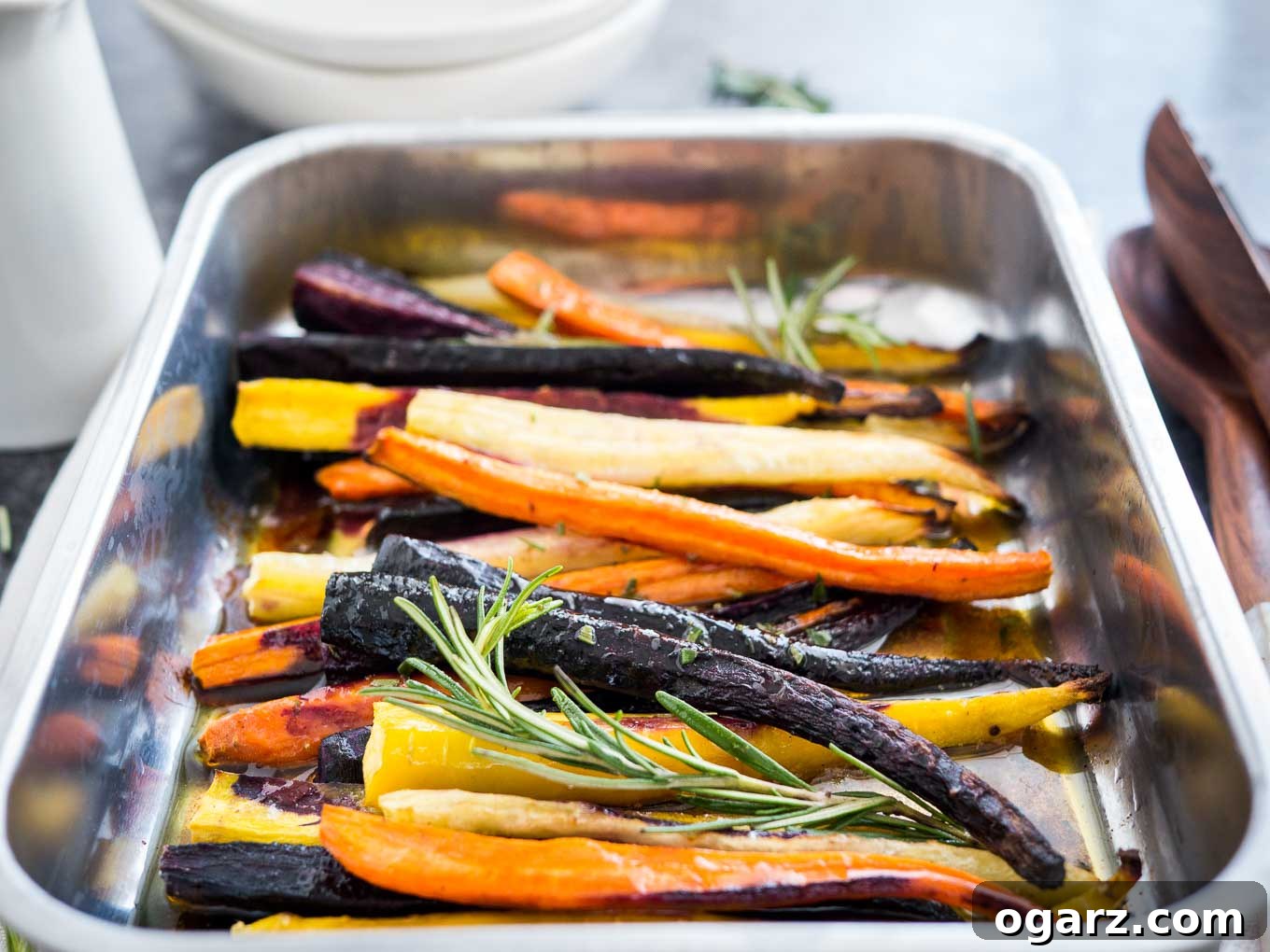 Brown Butter Maple Infused Roasted Rainbow Carrots 6 A roasting pan with roasted rainbow carrots garnished with sprigs of rosemary. There is a stack of plates in the background and salad tongs next to it.