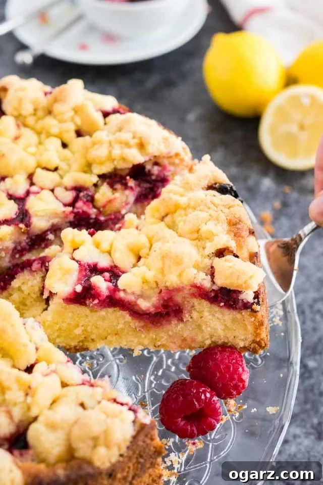 Effortless Lemon Raspberry Delight 3 A cake server lifting a perfectly baked slice of lemon raspberry cake with streusel topping from a elegant glass serving plate, garnished with fresh lemon slices and ripe raspberries.