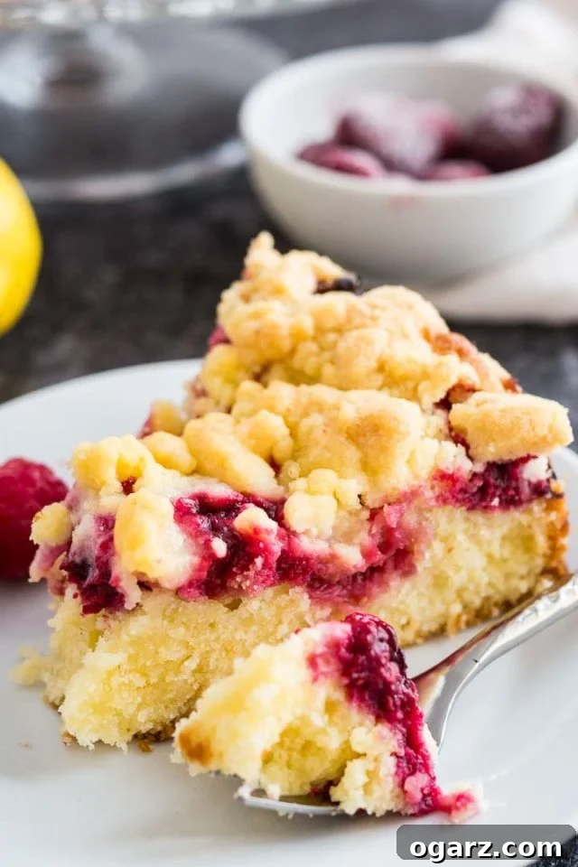 Effortless Lemon Raspberry Delight 4 A close-up shot of a single slice of lemon raspberry cake with streusel topping on a white plate, featuring a fork with a small piece of the cake, beautifully garnished with fresh lemon wedges and ripe raspberries.