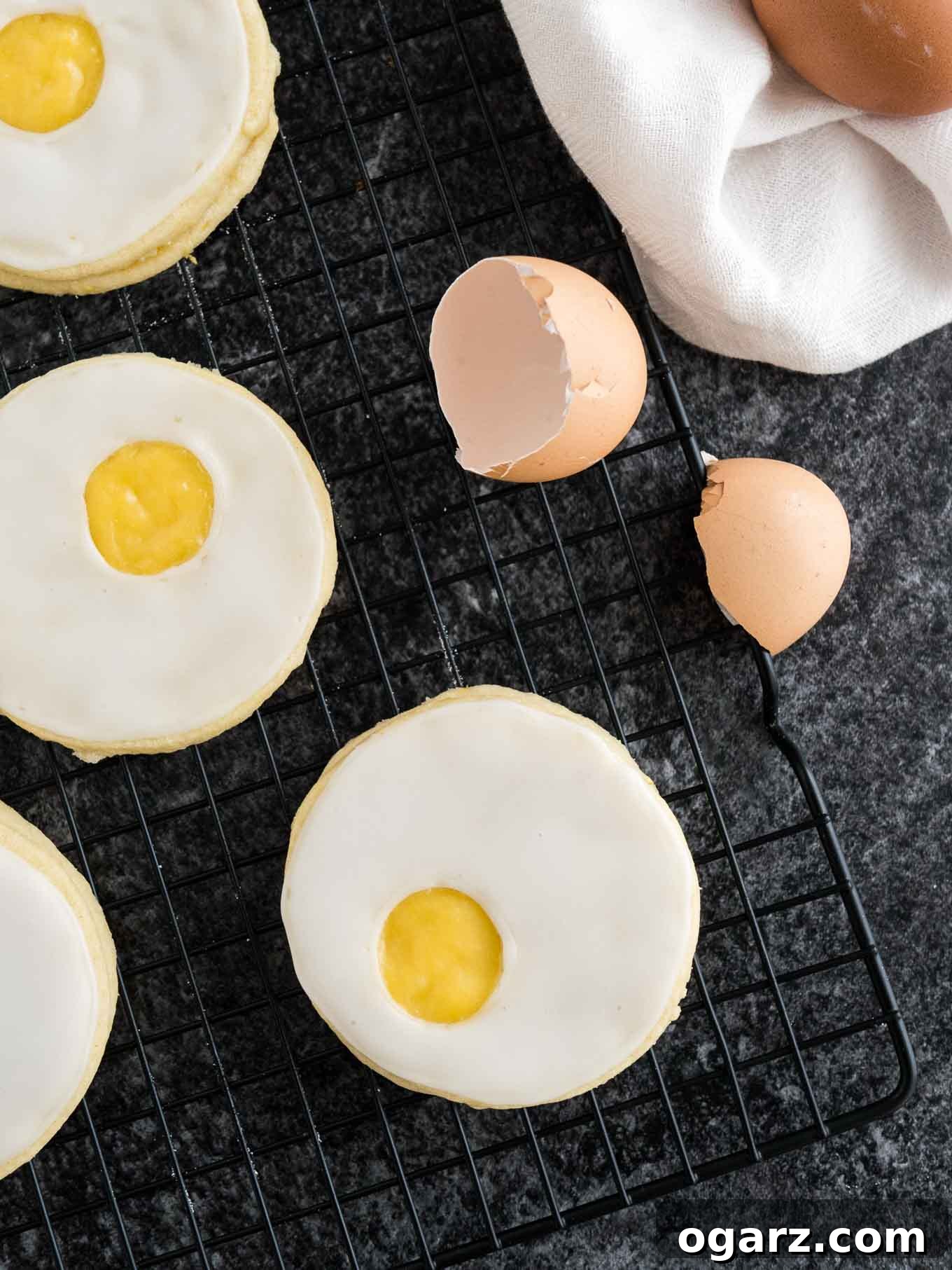 Sweet Easter Cookie Delights 3 A cooling rack holds several perfectly baked Easter sugar cookies, resembling sunny-side-up eggs with their lemon curd centers. Nearby, a scattering of eggshells and a soft white dishtowel complete the scene, suggesting fresh baking and a delightful spring treat.