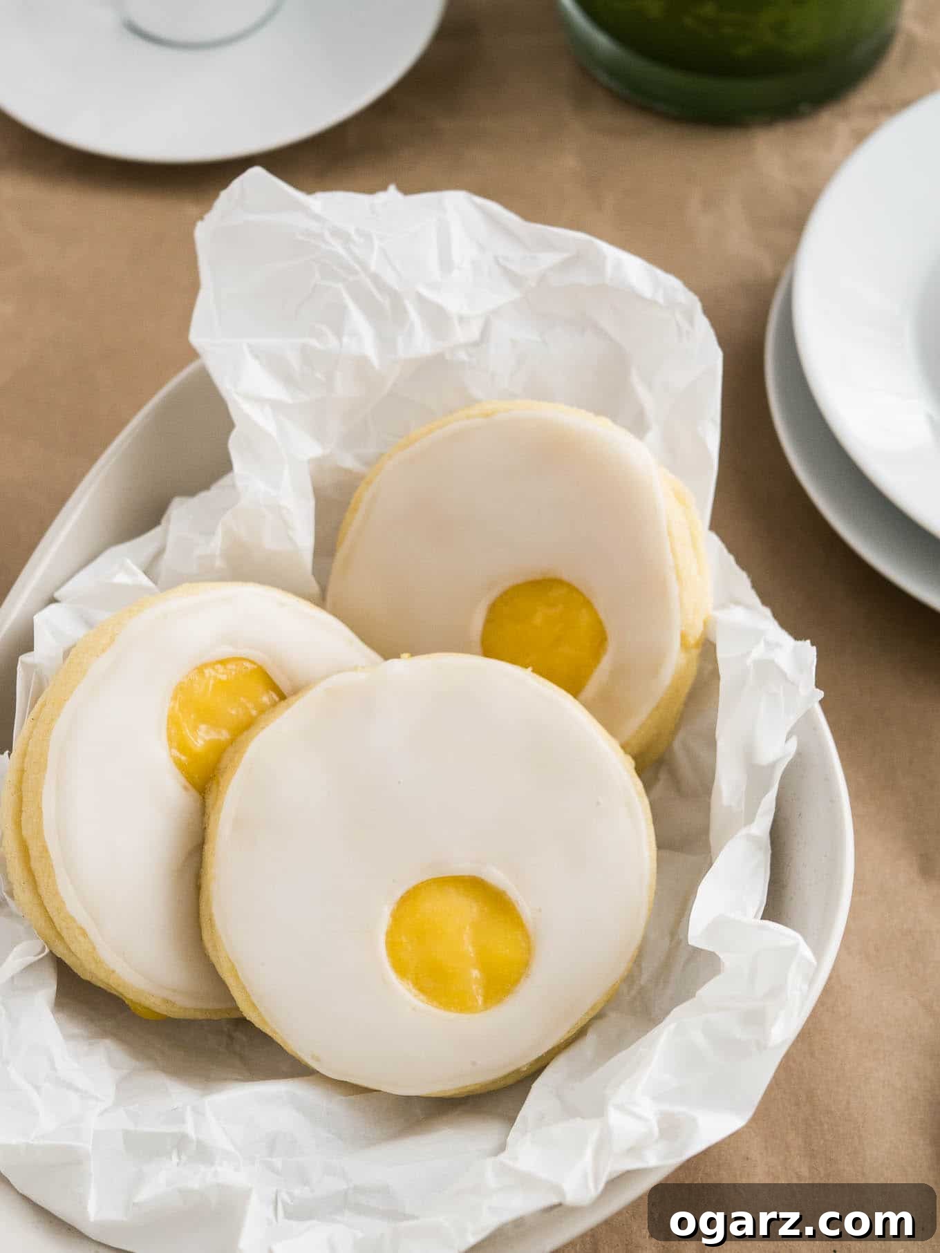 Sweet Easter Cookie Delights 4 Three beautiful Easter sugar cookies, resembling sunny-side-up eggs with their bright lemon curd centers, are neatly arranged in a bowl lined with brown parchment paper, positioned beside a stack of clean white plates. This charming display evokes a sense of homemade goodness and festive springtime baking.