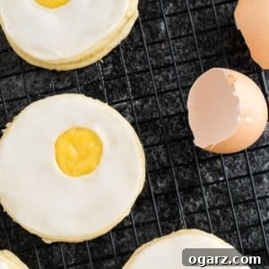 Several Easter sugar cookies with lemon curd, artfully resembling sunny-side-up eggs, are arranged on a black cooling rack. Eggshells scattered nearby provide a rustic touch, enhancing the theme of freshly baked springtime treats.