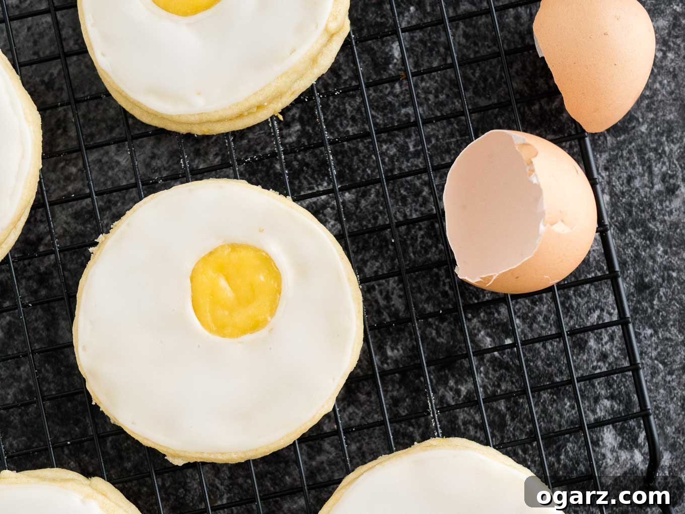 Sweet Easter Cookie Delights 6 A close-up shot of several delicate Easter sugar cookies, designed to mimic sunny-side-up eggs with their bright lemon curd filling. They are neatly arranged on a black cooling rack, accompanied by a few scattered eggshells, enhancing the theme of springtime baking and fresh, zesty treats.