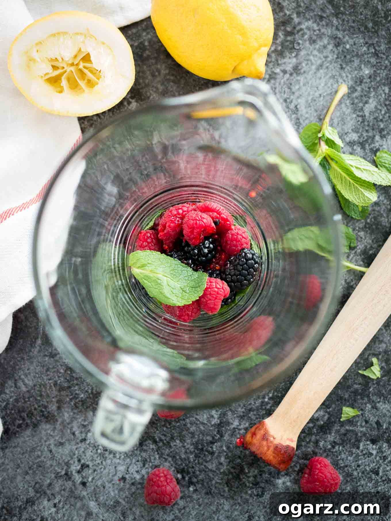 Craft Your Own Refreshing Raspberry Lemonade 3 Top-down shot of an empty glass jug with raspberries and blackberries and mint on a dark surface. There\'s a lime, a white and red dishtowel some mint leaves and a muddler next to it.
