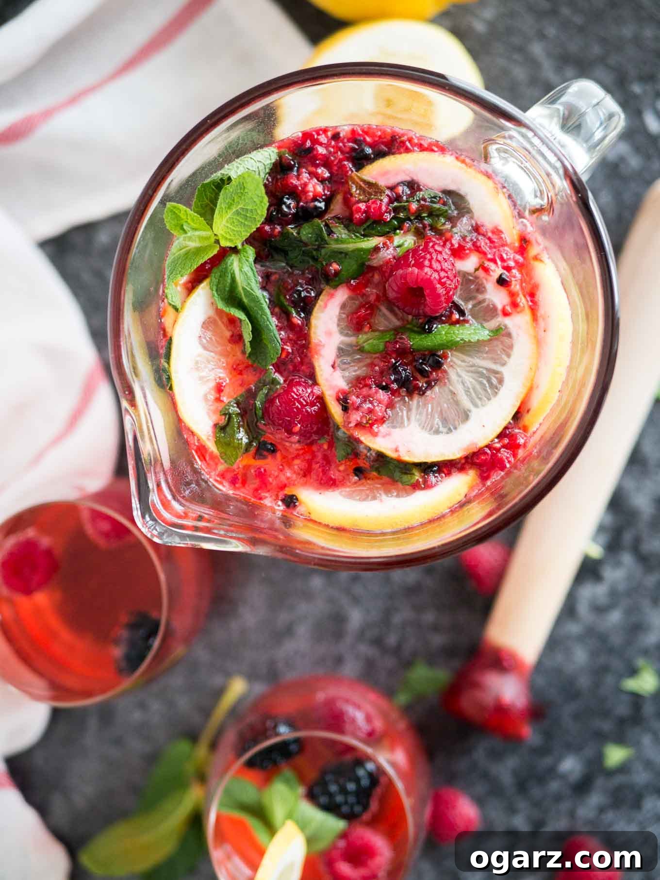 Craft Your Own Refreshing Raspberry Lemonade 4 Top-down shot of a glass pitcher with homemade raspberry lemonade with lemon slices, mint and raspberries and blackberries. There are two glasses of the lemonade, a muddler, mint leaves and a white and red dishtowel next to it.