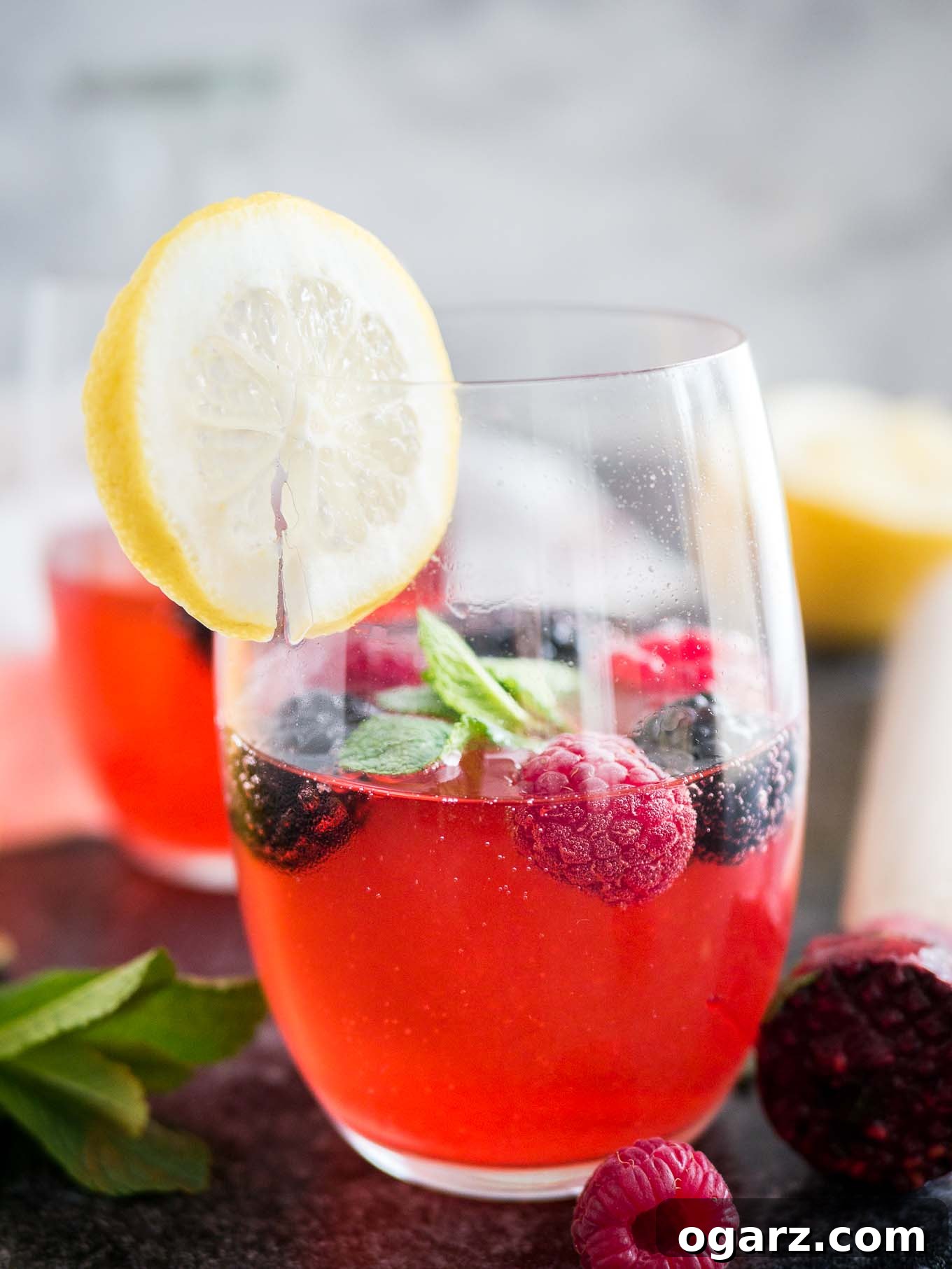 Craft Your Own Refreshing Raspberry Lemonade 5 Close-up of two glasses of raspberry lemonade with raspberries and mint in them and a slice of lemon on the rim. There\'s a muddler, mint and half a lemon next to it.