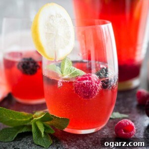 Craft Your Own Refreshing Raspberry Lemonade 6 Close-up of two glasses of raspberry lemonade with raspberries and mint in them and a slice of lemon on the rim. There's a jug with the rest of the lemonade in the background.