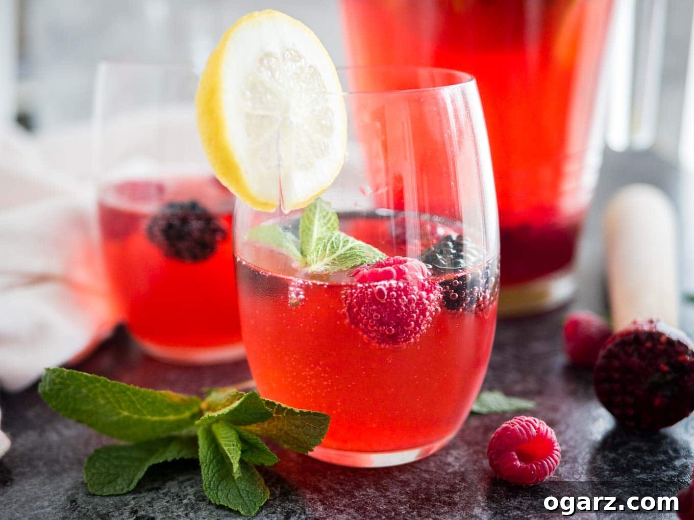 Craft Your Own Refreshing Raspberry Lemonade 7 Close-up of two glasses of raspberry lemonade with raspberries and mint in them and a slice of lemon on the rim. There\'s a jug with the rest of the lemonade in the background.