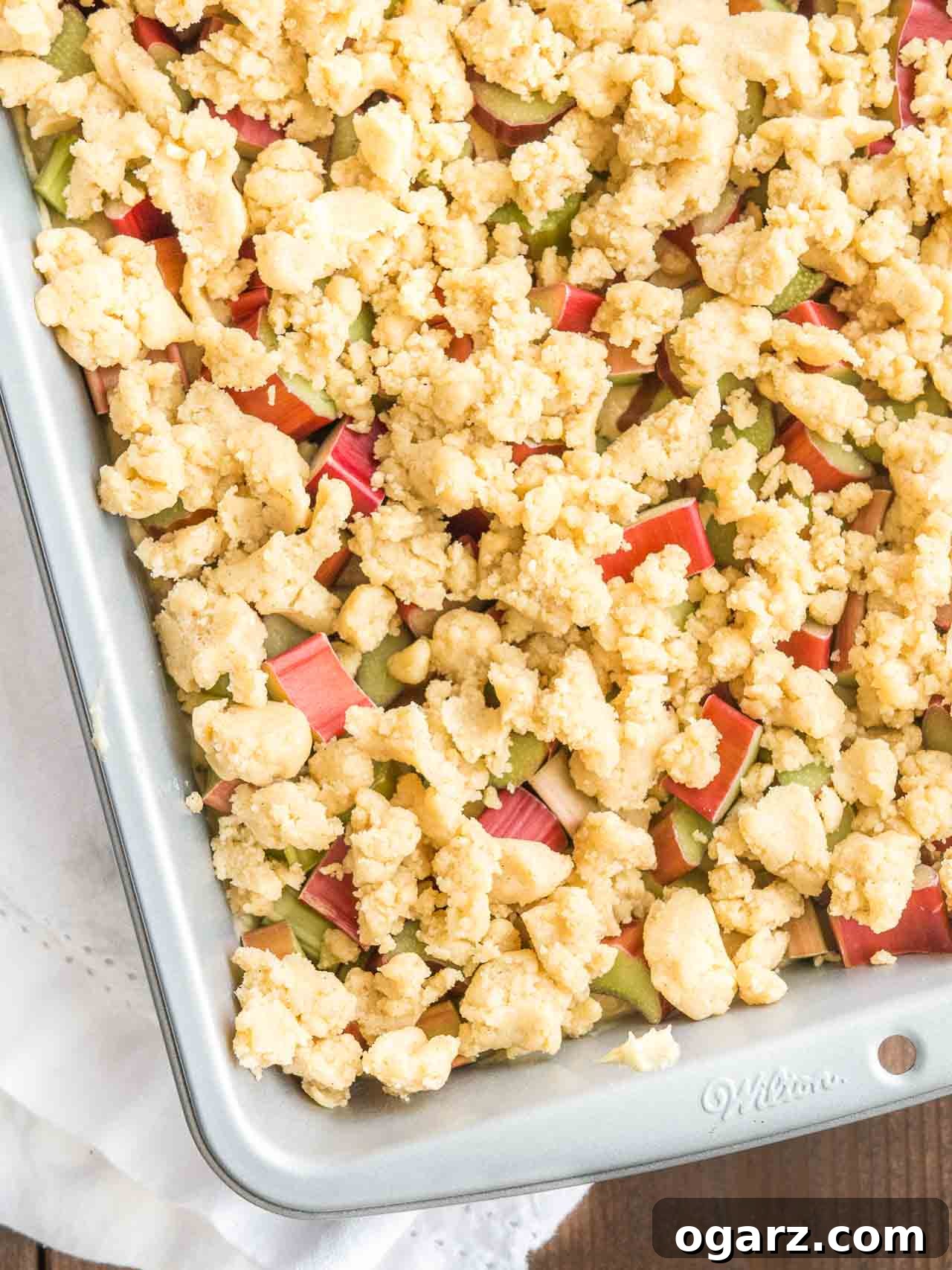 Tangy Rhubarb Streusel Cake 4 A grey baking pan filled with the unbaked Rhubarb Coffee Cake batter, evenly topped with chopped rhubarb and a generous layer of streusel, ready for the oven.