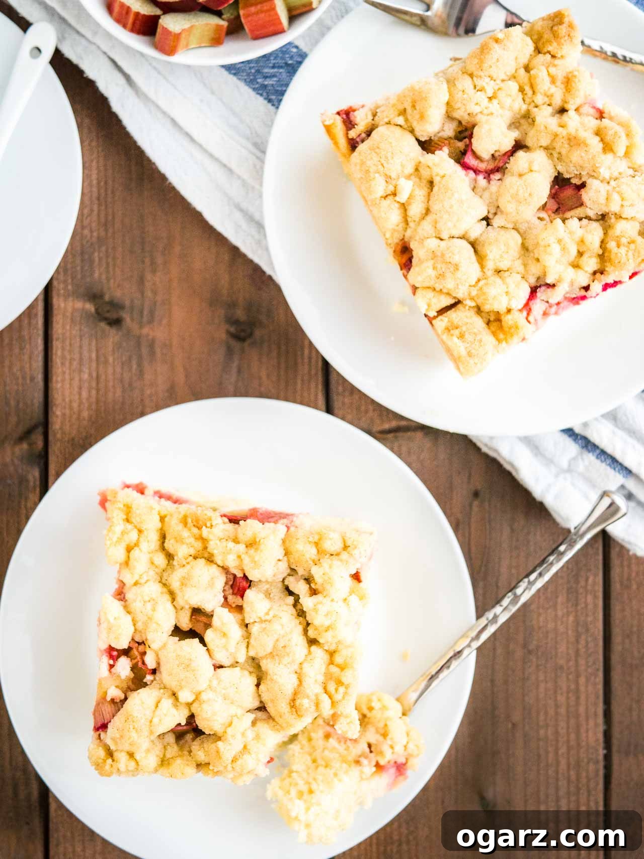 Tangy Rhubarb Streusel Cake 5 Top-down view of a delightful scene: a piece of Rhubarb Coffee Cake with streusel on a white plate, a fork having just removed a portion. Adjacent to it, another slice of cake, a small bowl of fresh rhubarb, and a warm cup of coffee complete the inviting tableau.