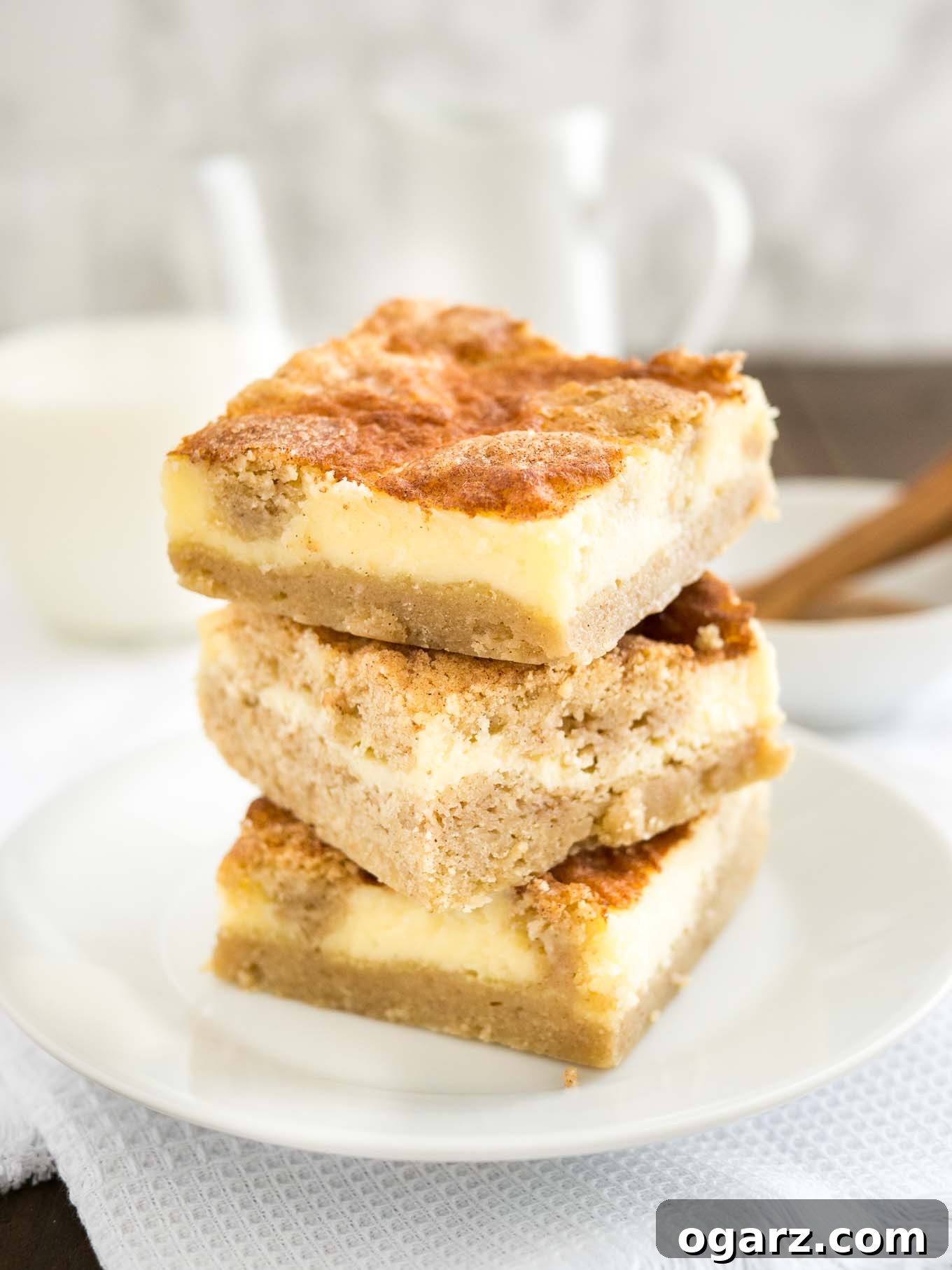 Cinnamon Sugar Snickerdoodle Cheesecake Bars 2 A stack of 3 snickerdoodle cheesecake bars on a white plate on a white tablecloth. There\'s a white bowl with cinnamon and a measuring spoon as well as a glass of milk in the background.