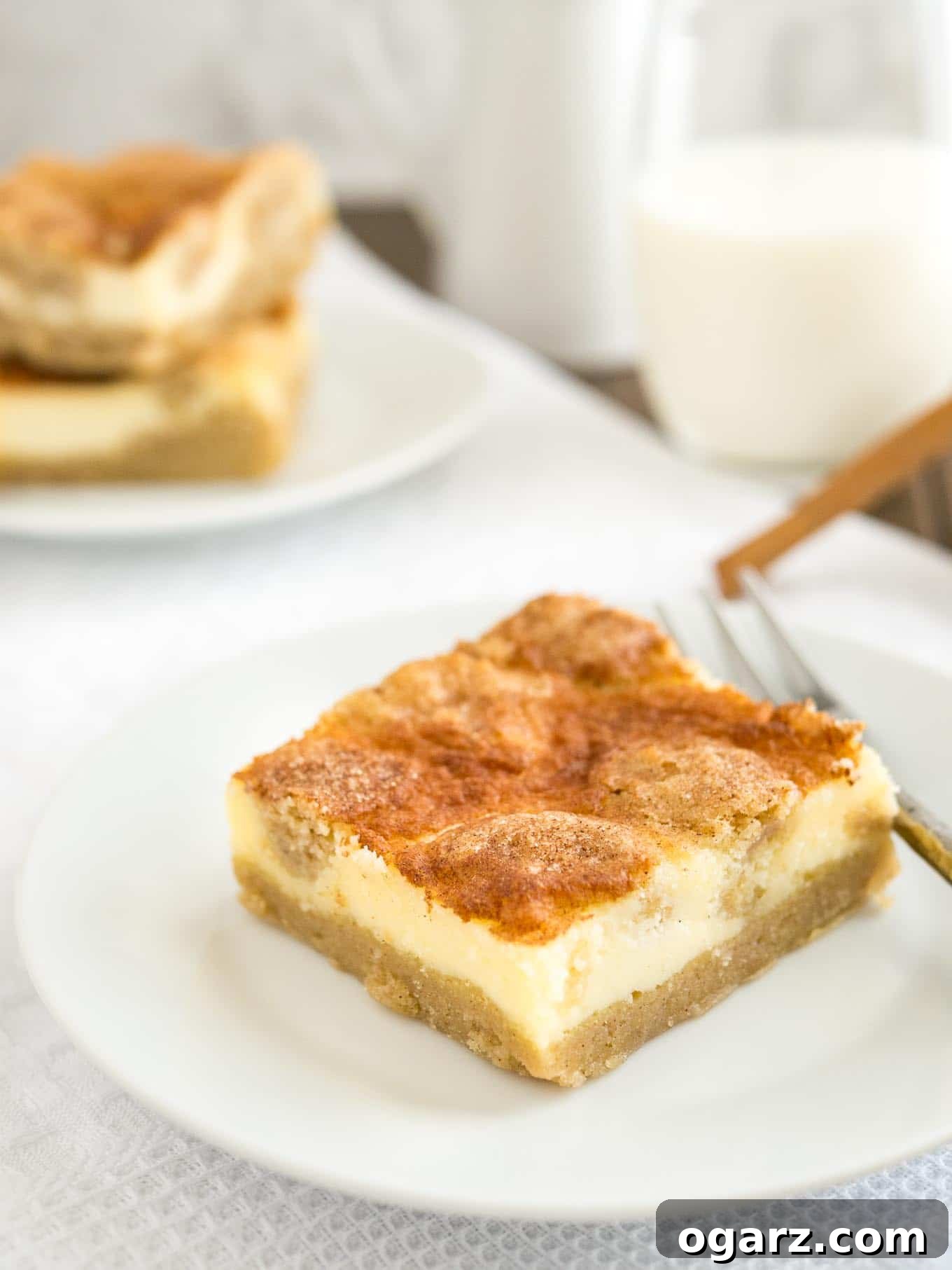 Cinnamon Sugar Snickerdoodle Cheesecake Bars 5 A white plate with a snickerdoodle cheesecake bar and a fork on it on a white tablecloth. There\'s a glass of milk and another plate with two cheesecake bars in the background, inviting you to take a bite.