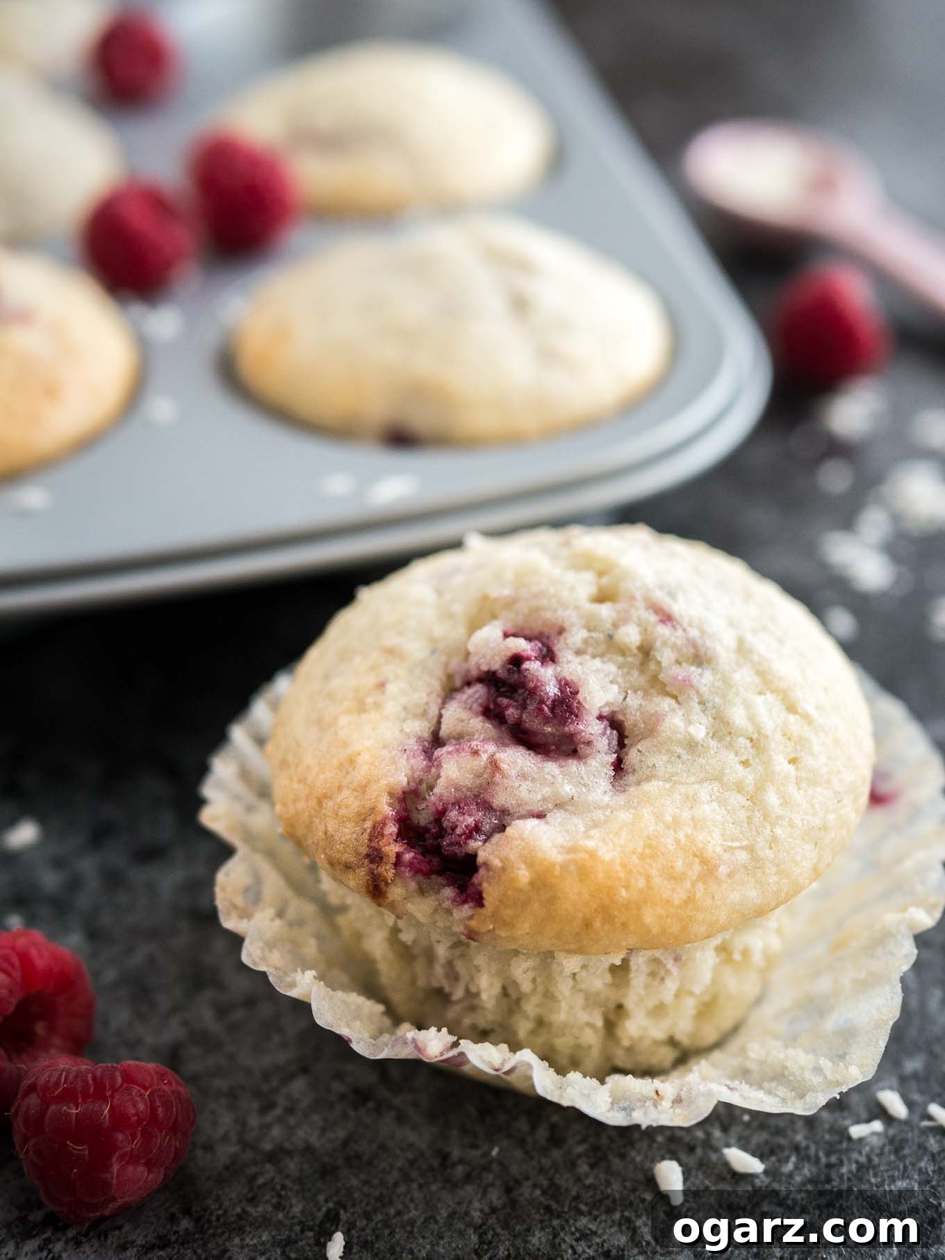 Tropical Raspberry Coconut Muffins 4 A coconut raspberry muffin with its liner opened sitting on a dark surface. There are raspberries, a pink measuring spoon with coconut flakes and a grey muffin pan with more muffins next to it.