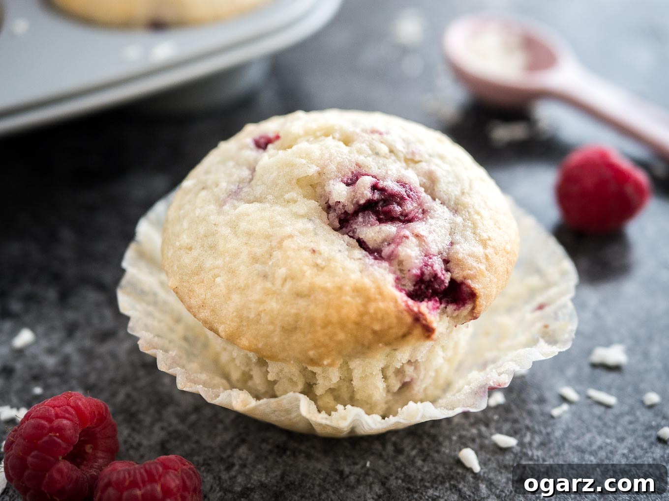 Tropical Raspberry Coconut Muffins 5 Close-up of a coconut raspberry muffin with its liner opened sitting on a dark surface. There are raspberries, a pink measuring spoon with coconut flakes and a grey muffin pan with more muffins next to it.