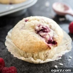 Close-up of a coconut raspberry muffin with its liner opened sitting on a dark surface. There are raspberries, a pink measuring spoon with coconut flakes and a grey muffin pan with more muffins next to it.