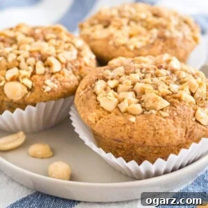 3 peanut butter banana muffins, topped with peanuts with their liners opened on a grey plate on a white and blue dishtowel.