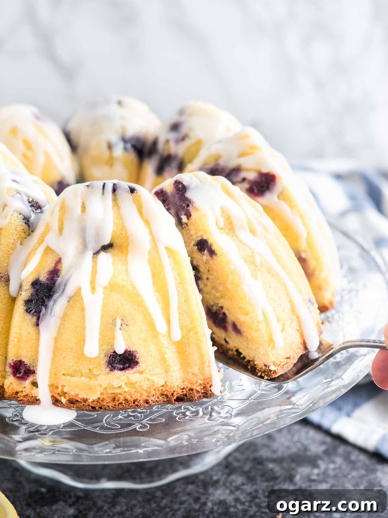 A stunning Lemon Blueberry Bundt Cake, beautifully drizzled with a vibrant lemon glaze, presented on an elegant glass serving platter. A cake server is positioned, ready to lift a delectable slice, while a charming white and blue dishtowel lies casually in the background, hinting at a homemade culinary creation.