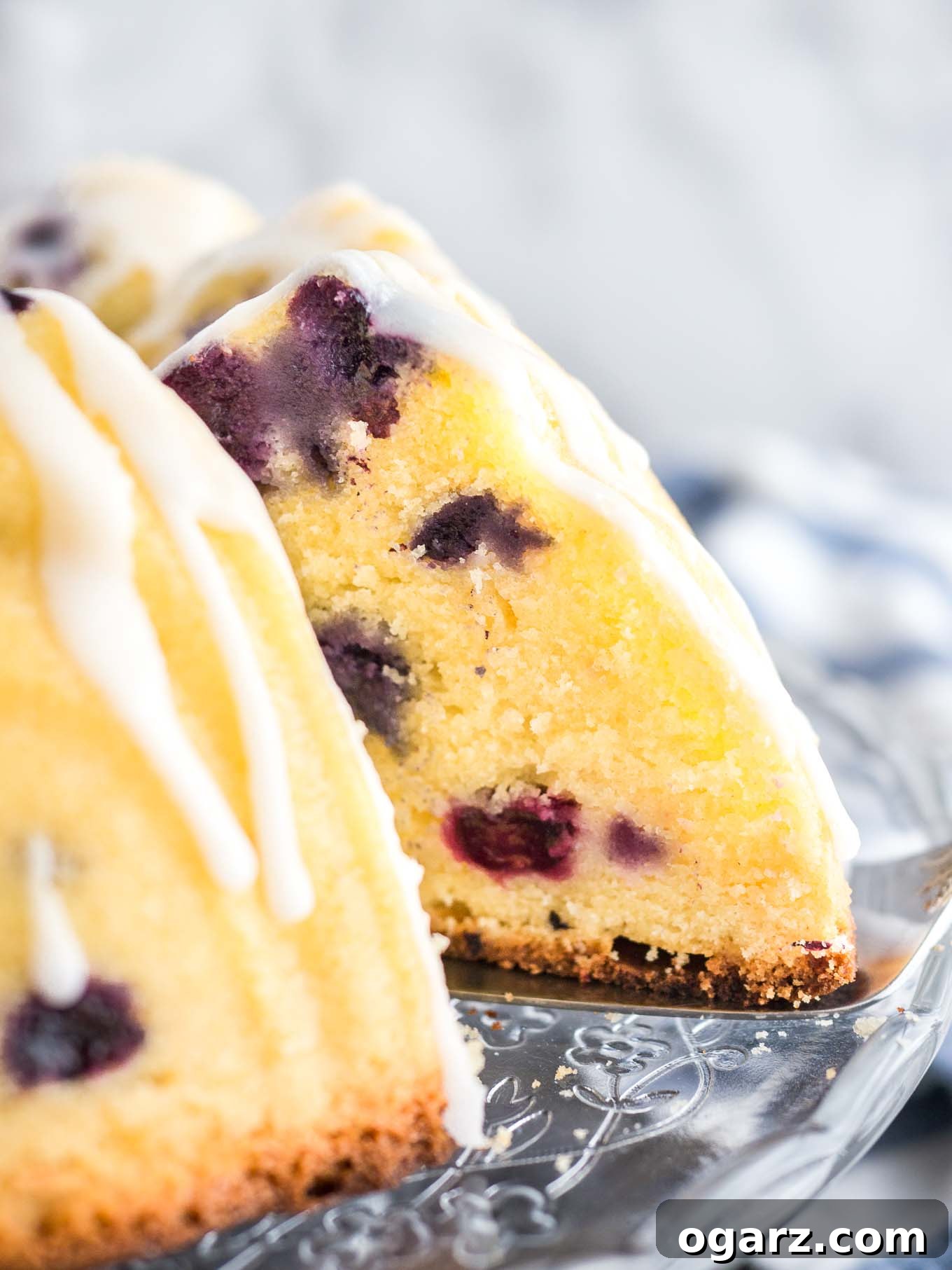 A close-up view of a magnificent Lemon Blueberry Bundt Cake on a glass serving platter, glistening with a fresh lemon glaze. A slice has been gracefully removed, revealing the moist crumb and scattered blueberries, with a cake server tucked underneath. A white and blue dishtowel adds a homely touch in the background.