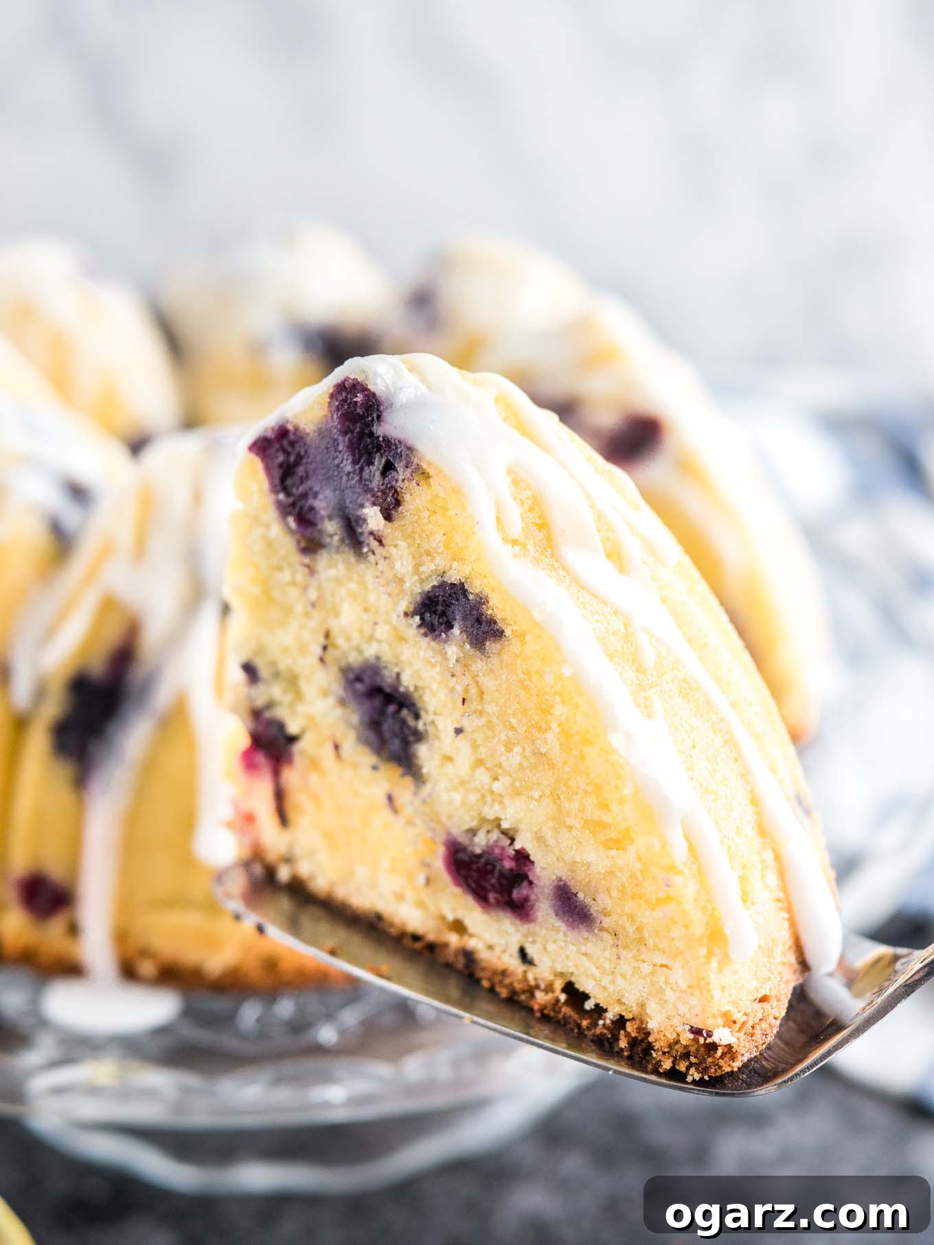 A delectable slice of Lemon Blueberry Bundt Cake delicately balanced on a cake server in the foreground. In the soft-focus background, the remaining beautifully glazed bundt cake sits on a glass serving platter, inviting another taste.