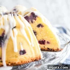 A lemon blueberry bundt cake on a glass serving platter with lemon glaze. A slice has been removed and a cake server is wedged under the cake. There's a white and blue dishtowel in the background.