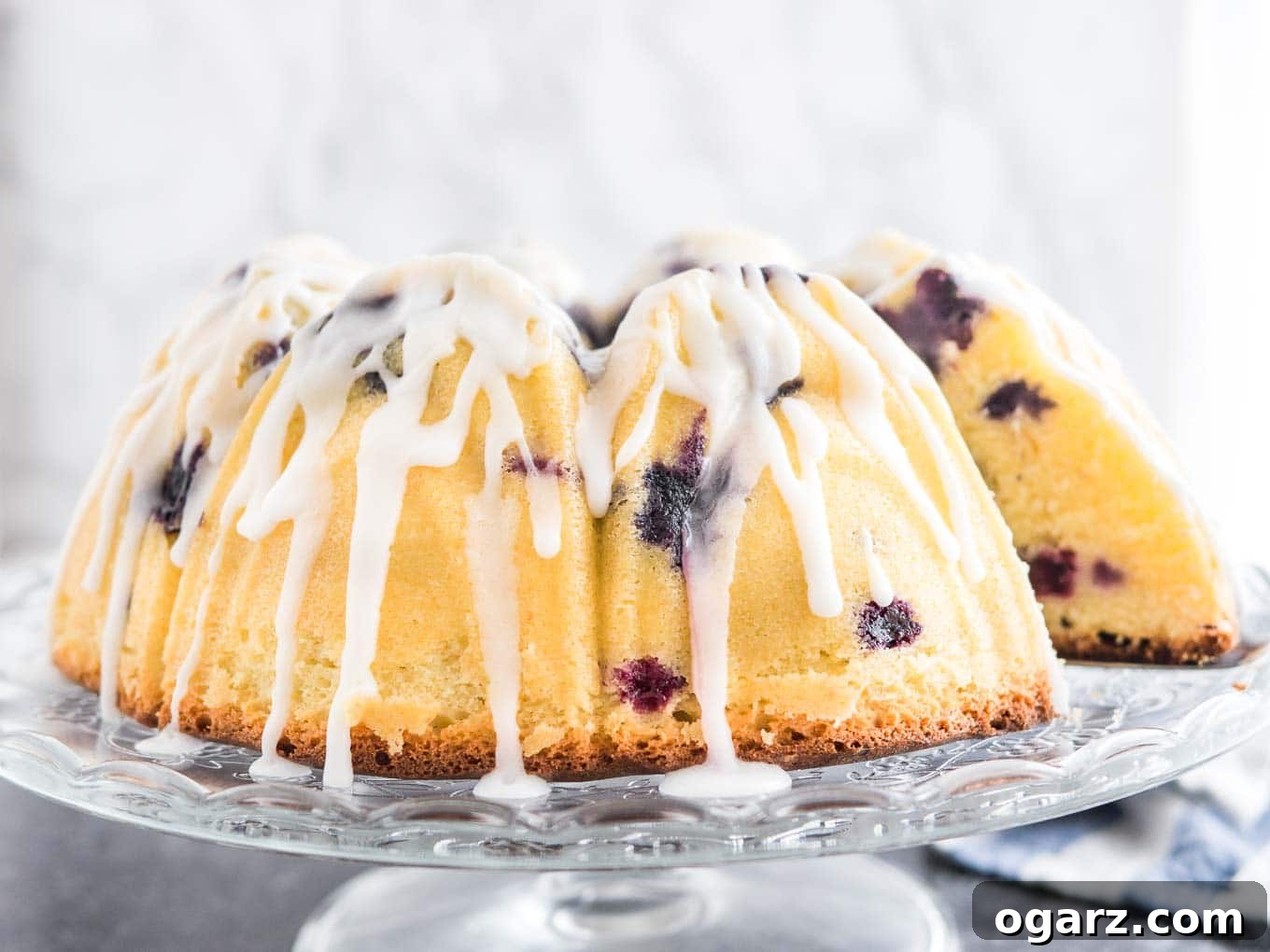 A beautifully glazed Lemon Blueberry Bundt Cake sits gracefully on a glass serving platter. A slice has been removed, revealing the soft, fruit-studded interior, while a white and blue dishtowel subtly enhances the rustic, homemade charm in the background.