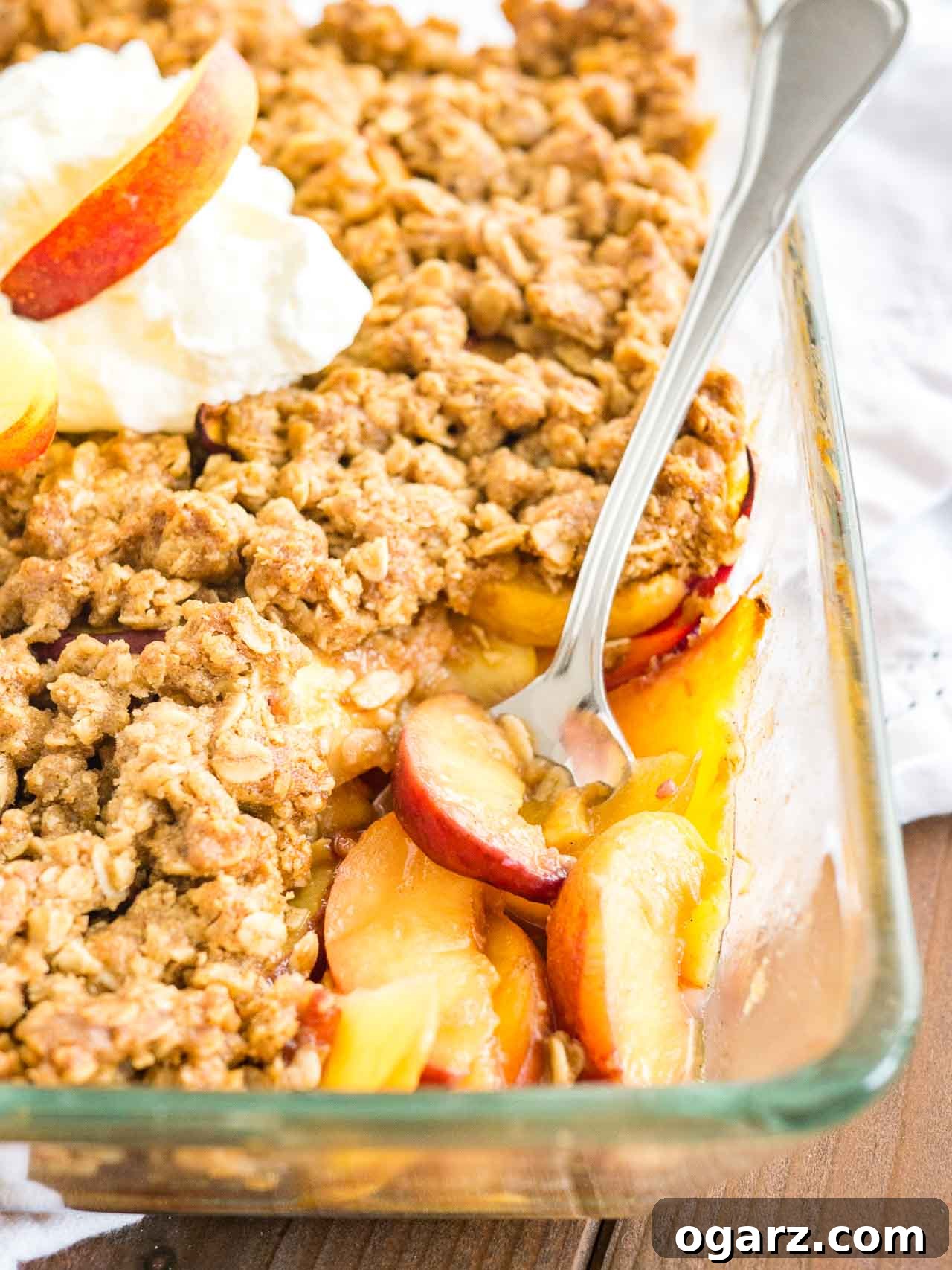 A glass baking dish with a layer of peaches topped by a layer of crisp on a white dishtowel, garnished with whipped cream and peaches.