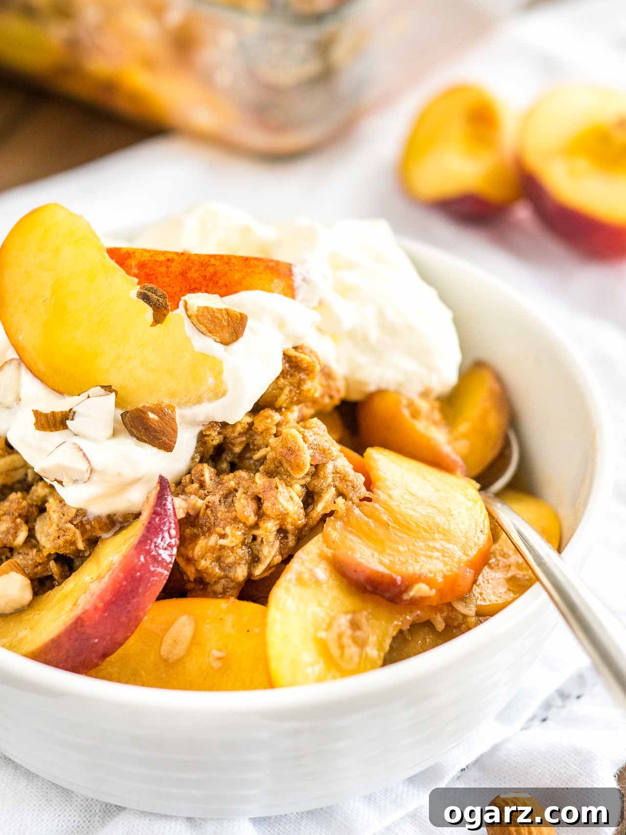 A white bowl of peach crisp with almonds and whipped cream on a white dishtowel with peach wedges in the back. A spoon is digging into the crisp.