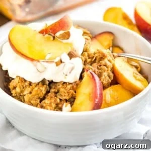 A white bowl of peach crisp with almonds and whipped cream on a white dishtowel with peach wedges in the back.