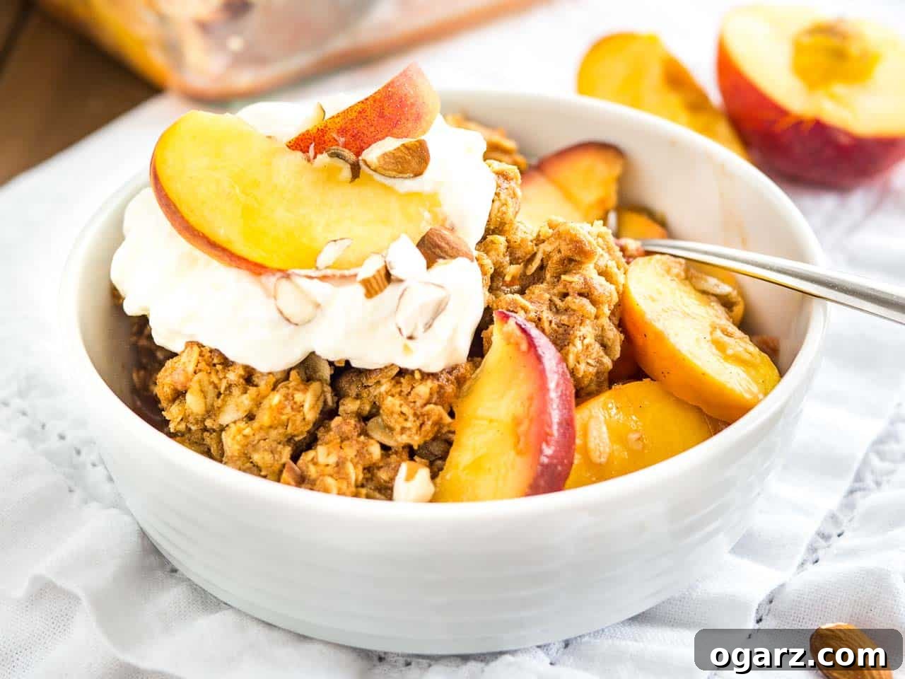 A white bowl of peach crisp with almonds and whipped cream on a white dishtowel with peach wedges in the back.