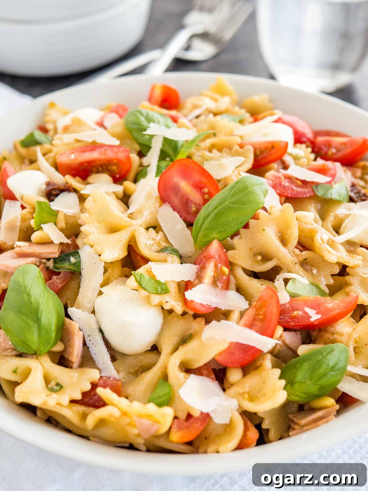 Zesty Italian Pasta Salad 2 Close-up of a white bowl with pasta salad with Italian dressing, with tomato, mozzarella, basil leaves and shavings of Parmiggiano. The bowl is sitting on a white dishtowel with forks, a glass of water and white bowls.