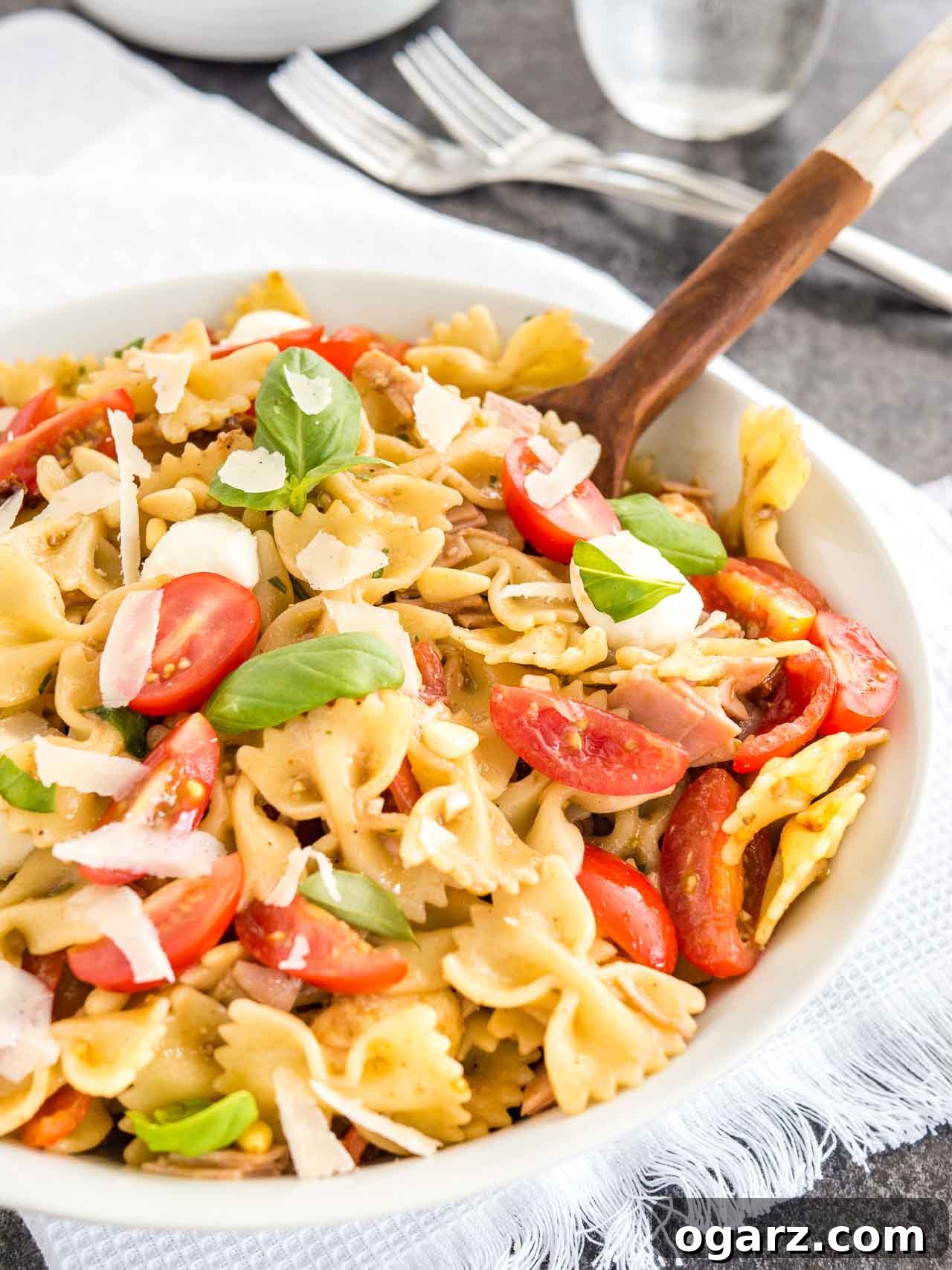 Zesty Italian Pasta Salad 3 A white bowl with pasta salad with italian dressing, with tomato, mozzarella, basil leaves and shavings of Parmiggiano with a salad tong in it. The bowl is sitting on a white dishtowel with forks next to it.