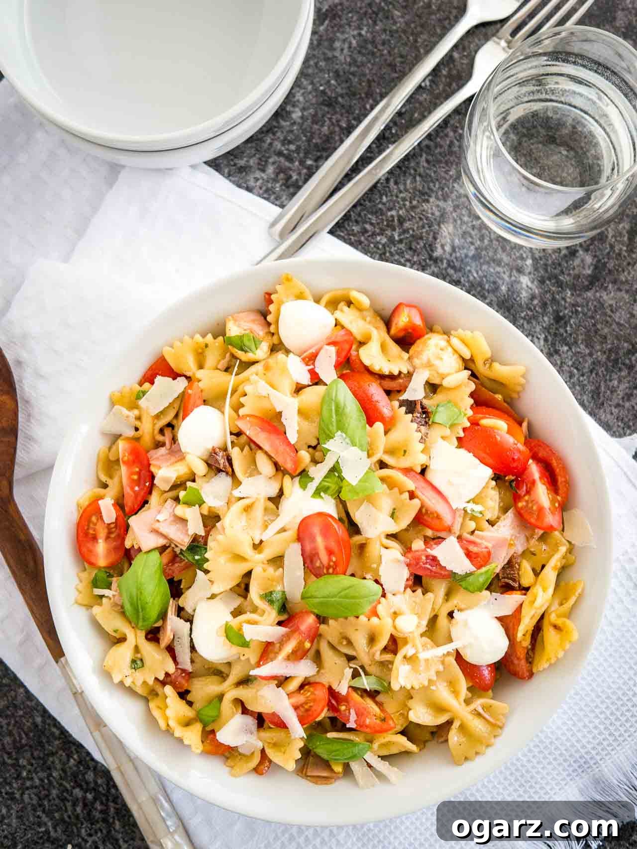 Zesty Italian Pasta Salad 4 Top-down shot of a white bowl with pasta salad with italian dressing, with tomato, mozzarella, basil leaves and shavings of Parmiggiano. The bowl is sitting on a white dishtowel with forks, a glass of water and salad tongs next to it.
