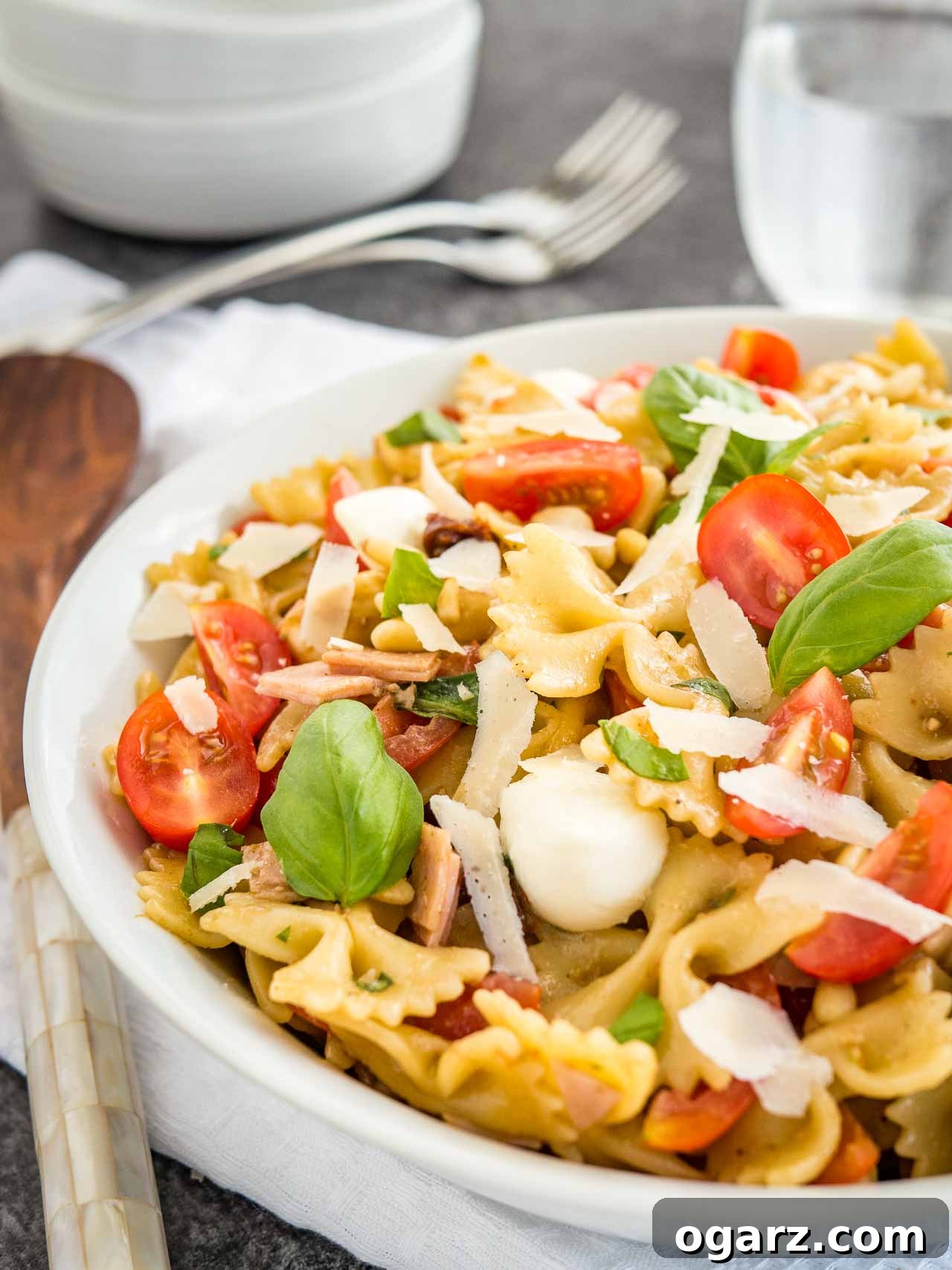 Zesty Italian Pasta Salad 5 Close-up of a white bowl with pasta salad with Italian dressing, with tomato, mozzarella, basil leaves and shavings of Parmiggiano. The bowl is sitting on a white dishtowel with forks, a glass of water and salad tongs in the background.