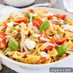 A white bowl with pasta salad with italian dressing, with tomato, mozzarella, basil leaves and shavings of Parmiggiano. The bowl is sitting on a white dishtowel with forks, a glass of water and salad tongs in the background.
